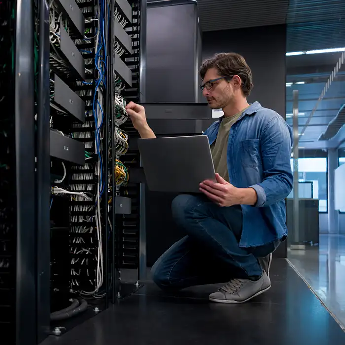 man kneeling with laptop in front of network cables