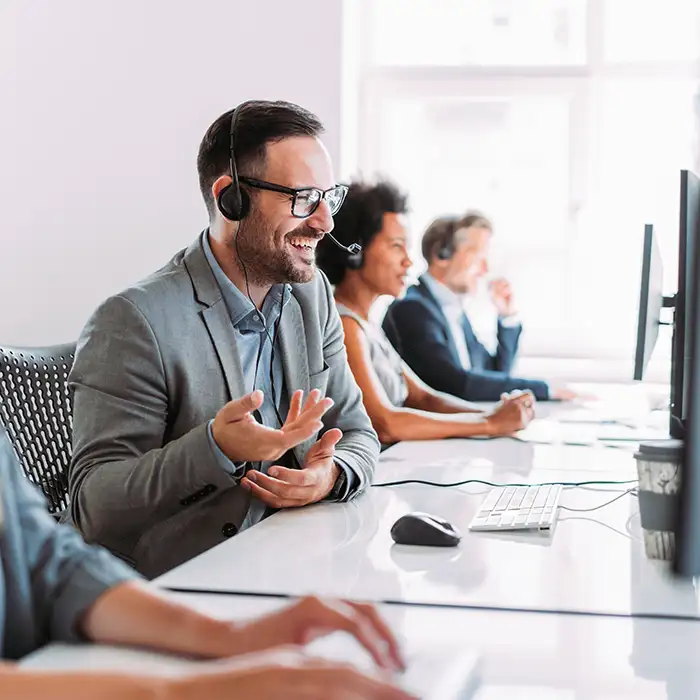 man with headset smiling and looking at computer in office with other workers